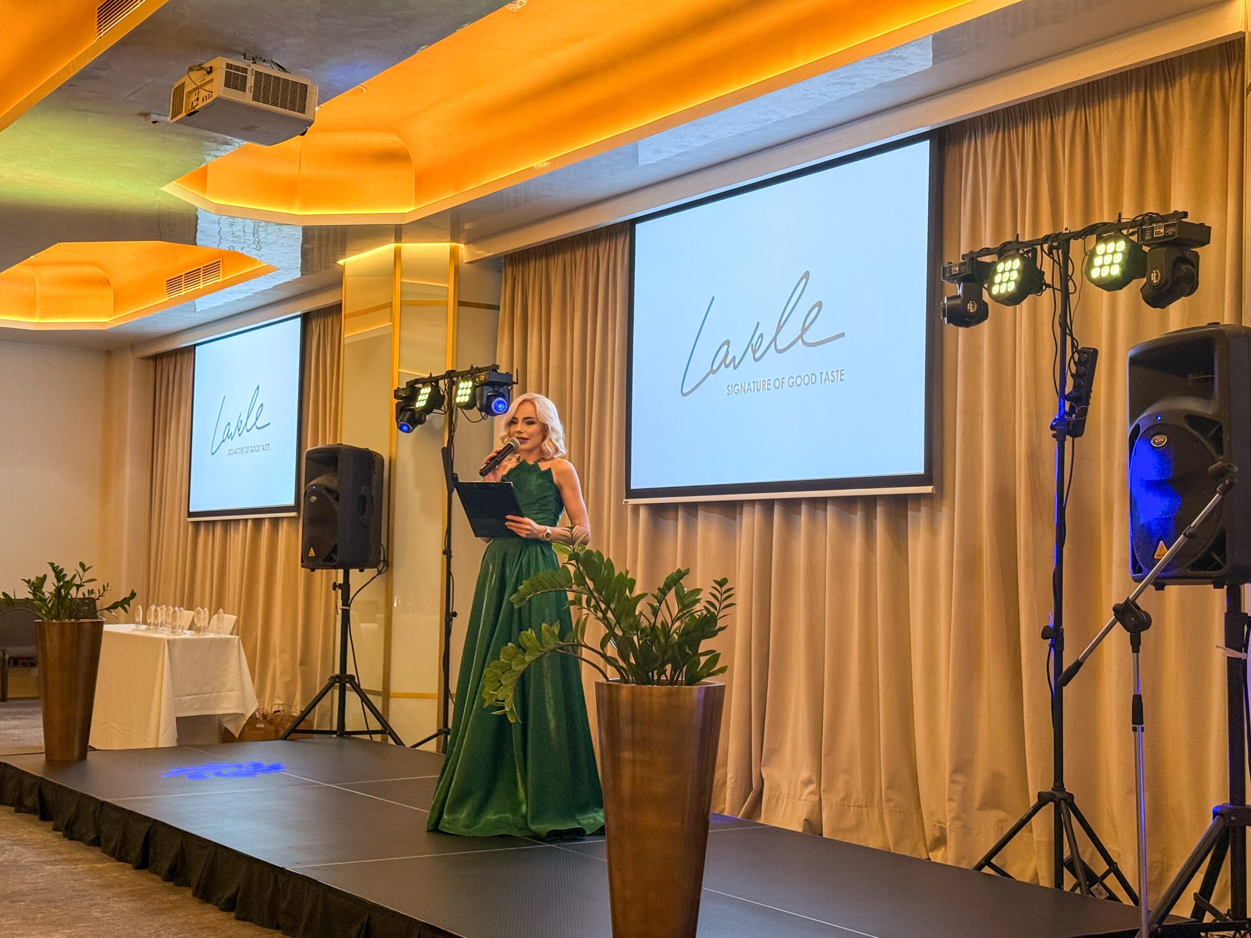 Female singer in a green gown performs on a stage with a handheld mic, two screens behind showing a logo, and warm orange lighting behind her.