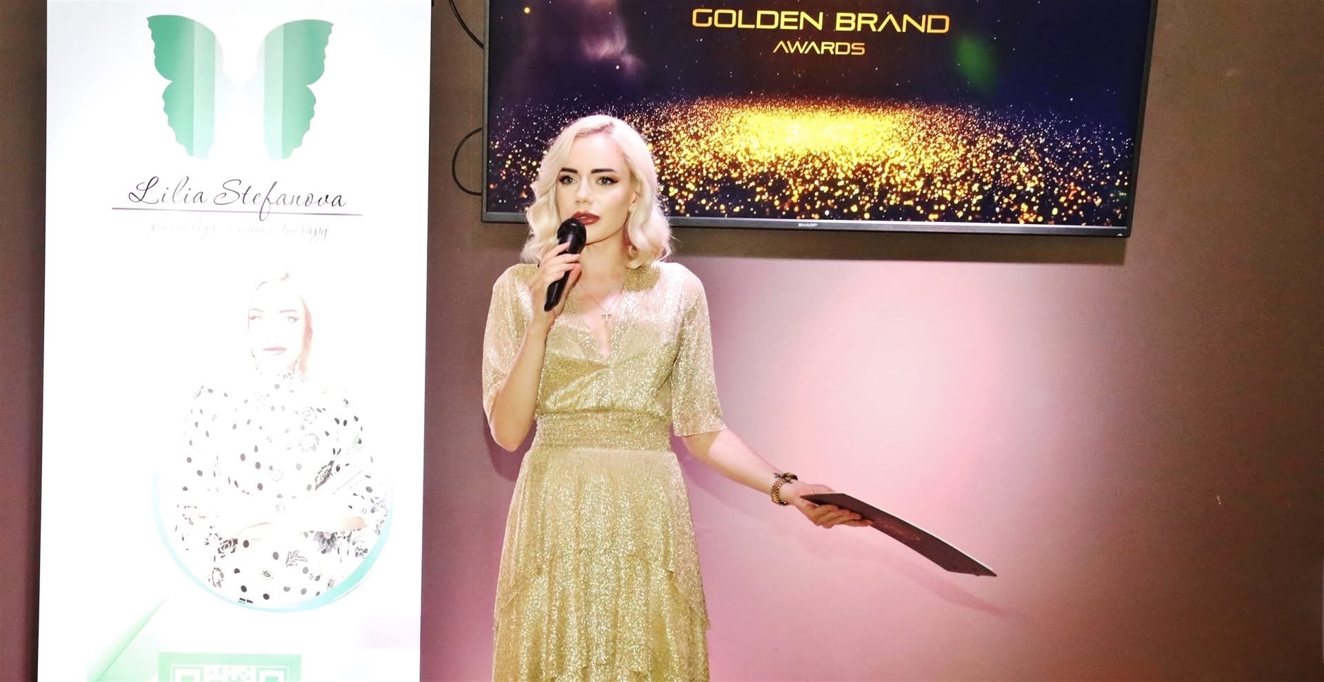 Female presenter in a glittery gold dress speaks into a handheld microphone on stage at the Golden Brand Awards.
