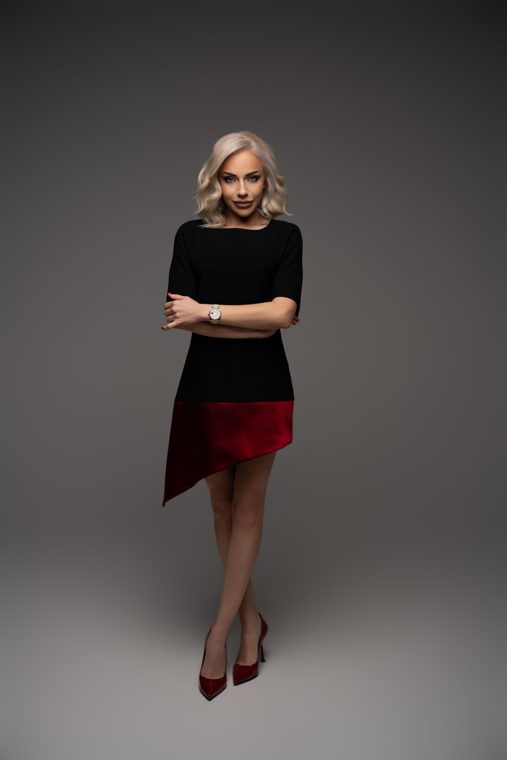 Portrait of a stylish blonde woman in a black dress with a red hem, arms crossed, wearing a gold watch, and red heels against a gray backdrop.
