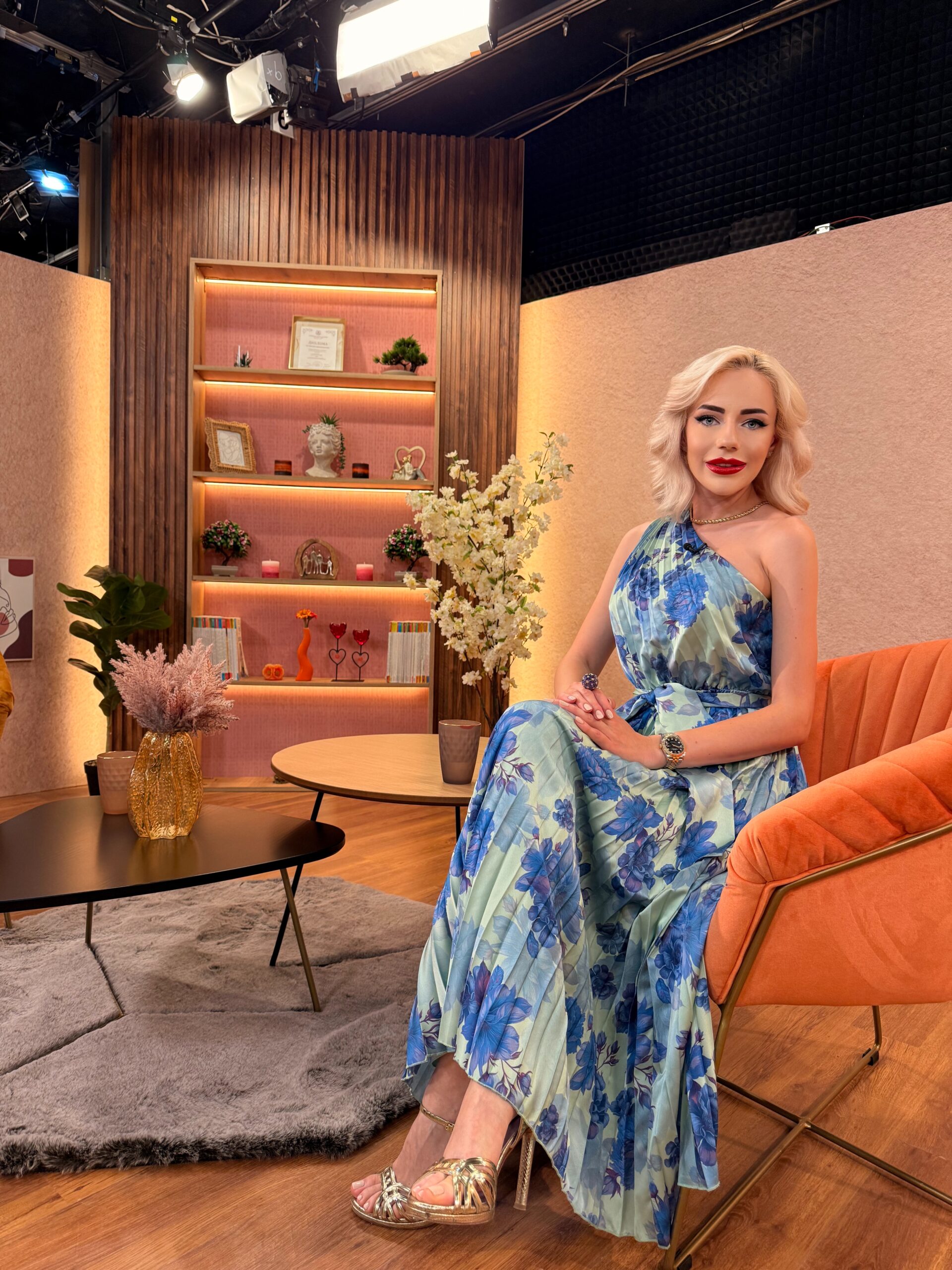 Woman with blonde hair in a blue floral dress sits on an orange chair in a studio set with shelves and warm lighting behind her, looking at the camera.